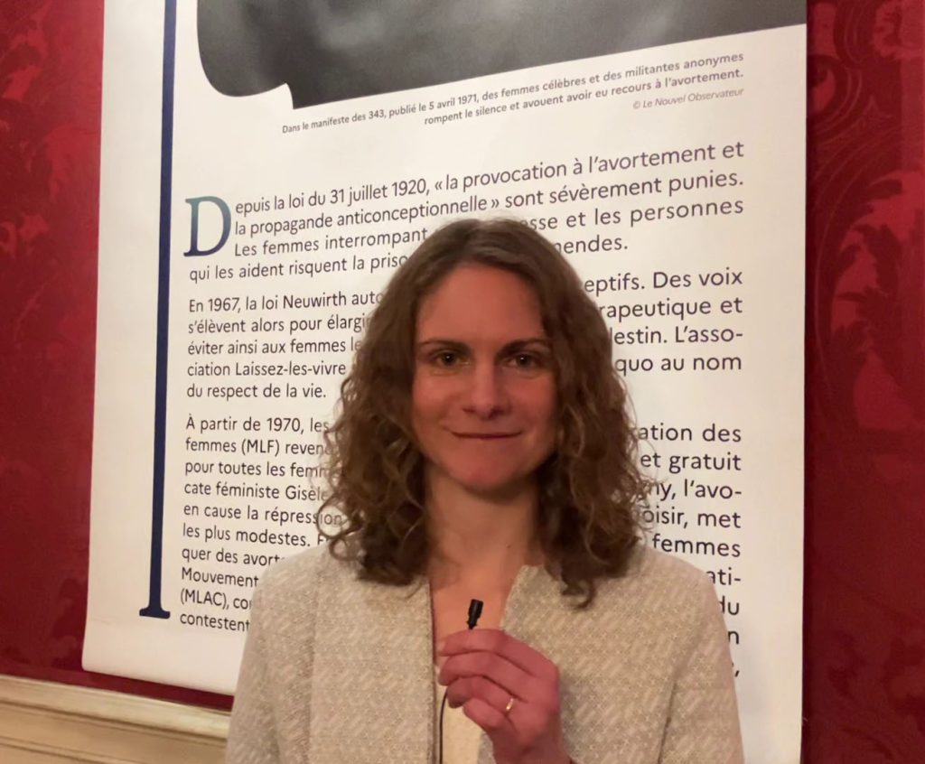 Photo de Charlène Fanchon posant devant un panneau de l'exposition Loi sur l'IVG 1974, aux Archives nationales en mars 2024.